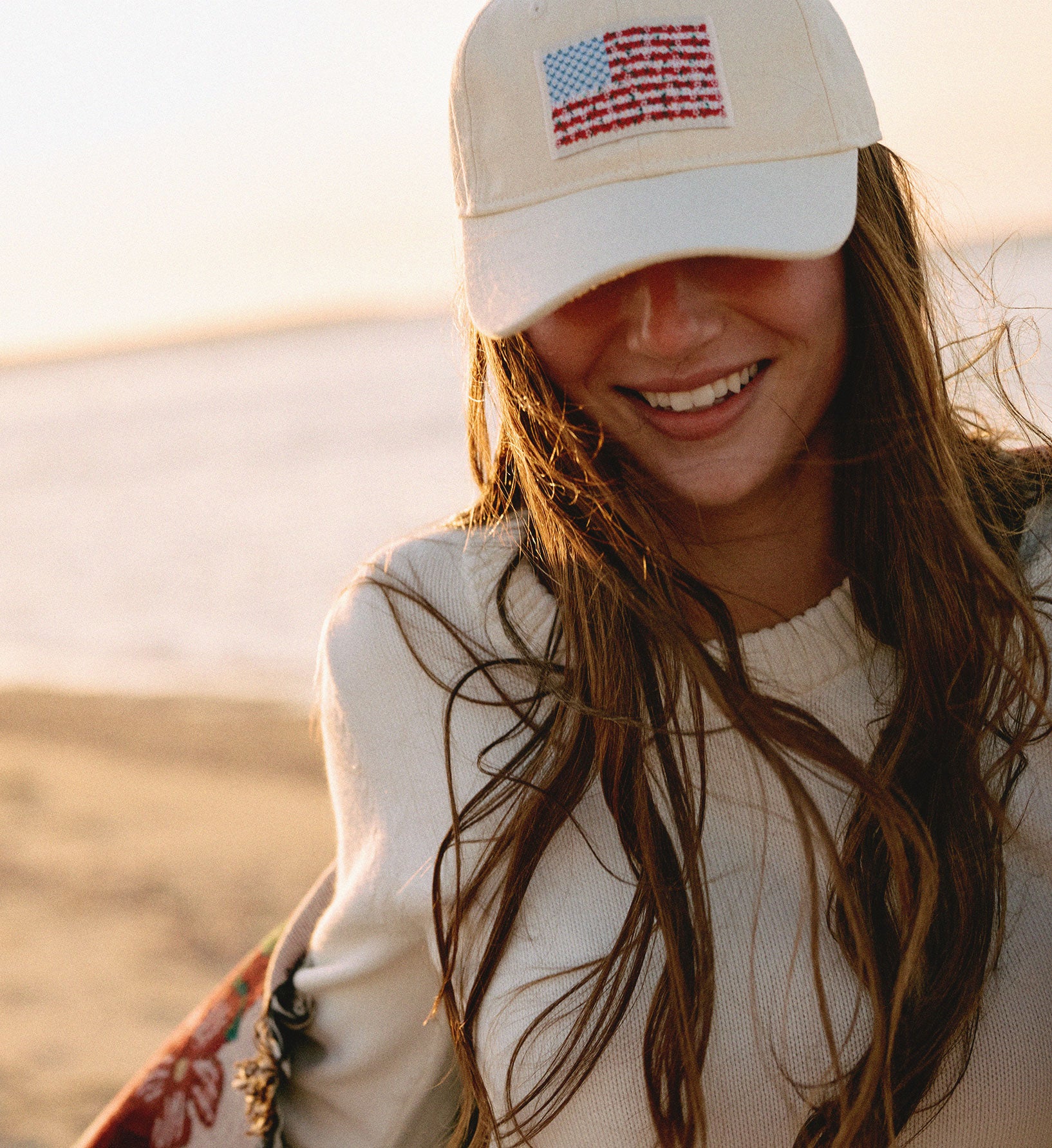 Woman wearing a beige cap with an American flag design, standing outdoors with a blurred natural background.
